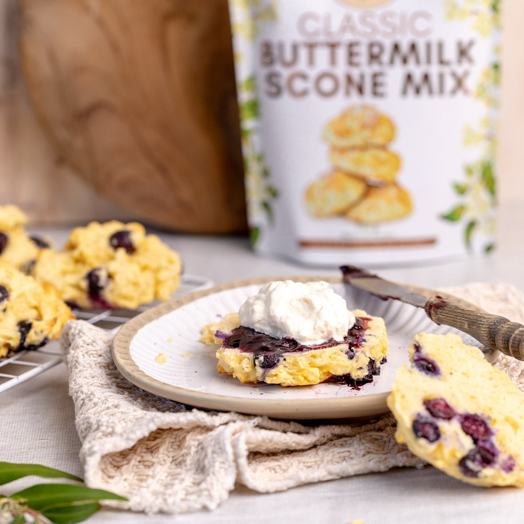 Secret Kiwi KitchenButtermilk scone with jam and cream on a plate, with a can of scone mix in the background.
