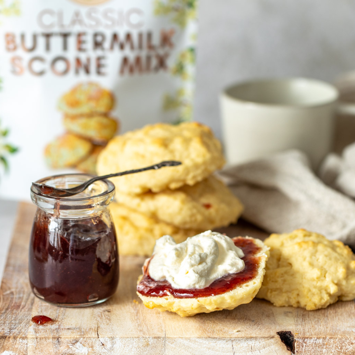 A glass jar of dark red jam sits beside a stack of golden flaky scones on a wooden board, with one scone split open and topped with whipped cream and jam in the foreground. A blurred package labeled Secret Kiwi Kitchen Classic Buttermilk Scone Mix is visible in the background, along with a mug and cloth.