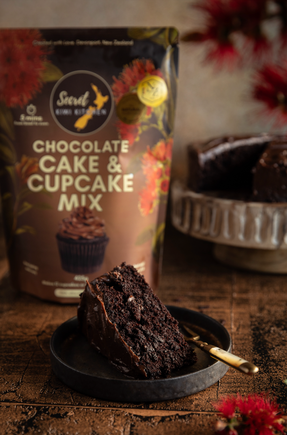 Secret Kiwi Kitchen Cake and Cupcake Mix package with a piece of chocolate cake on a black plate in front of it.
