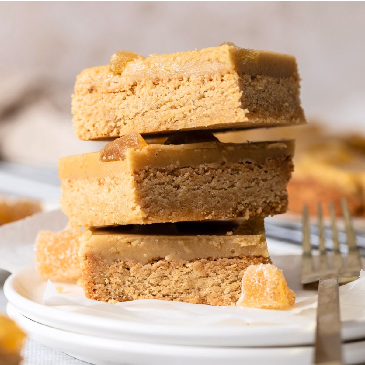 Three stacked square Secret Kiwi Kitchen Ginger Crunch Slice bars with a ginger icing on a white plate, with a fork and blurred gingerbread pieces in the background.