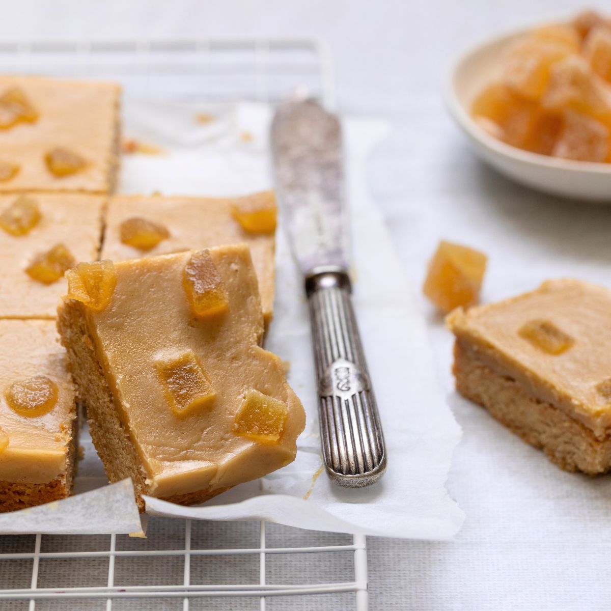 Secret Kiwi Kitchen ginger-frosted square dessert bars topped with candied ginger pieces, arranged on parchment paper on a cooling rack with a vintage knife, alongside a small bowl of additional fruit pieces.