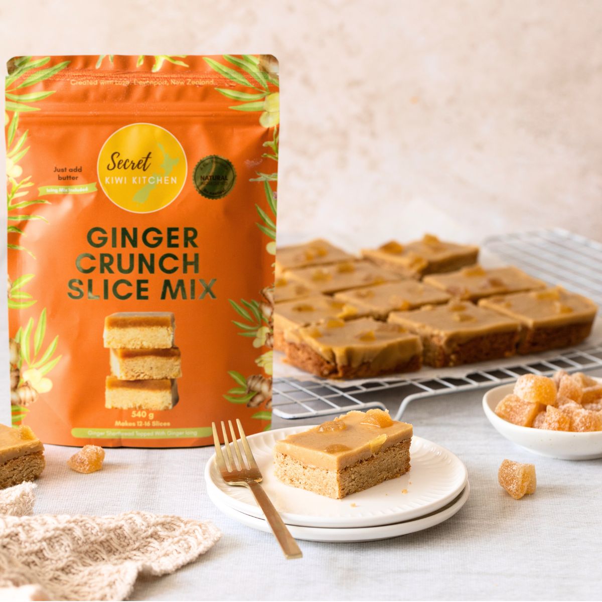 A stand-up pouch of Secret Kiwi Kitchen Ginger Crunch Slice Mix stands on a light surface beside plated gingerbread bars, a fork, and small pieces of candied ginger topping. The package is bright orange with green ginger palnt graphics and the product name in large black text.