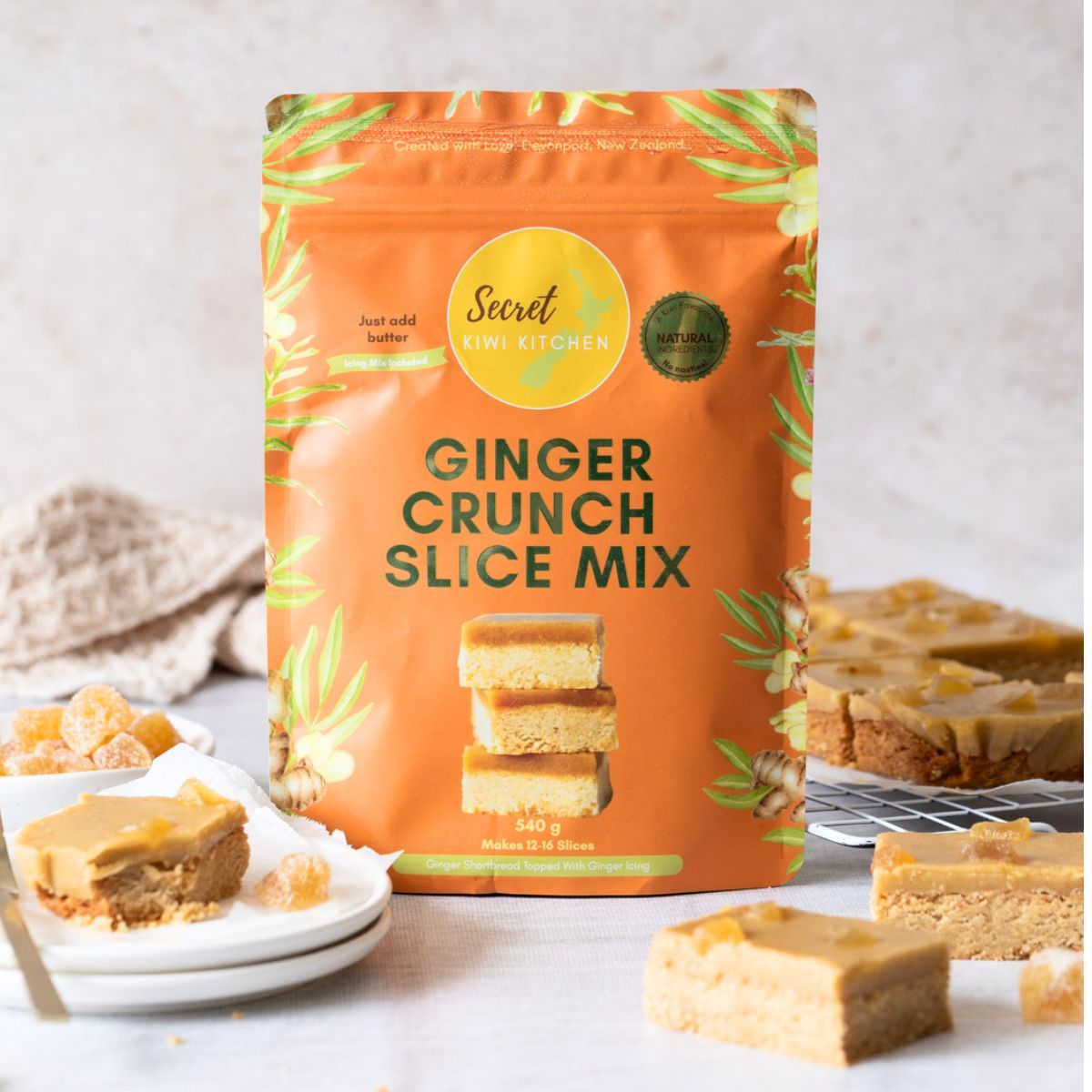 A stand-up pouch of Secret Kiwi Kitchen Ginger Crunch Slice Mix is shown on a light marble surface with several ginger bars and dried ginger pieces arranged around it.