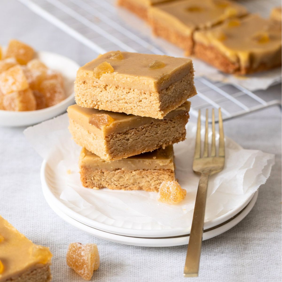 Three stacked square slices of Secret Kiwi Kitchen Ginger Crunch Slice bars topped with pieces of candied ginger, served on a white plate with a gold fork beside it. More slices and a cooling rack appear blurred in the background on a light tabletop.