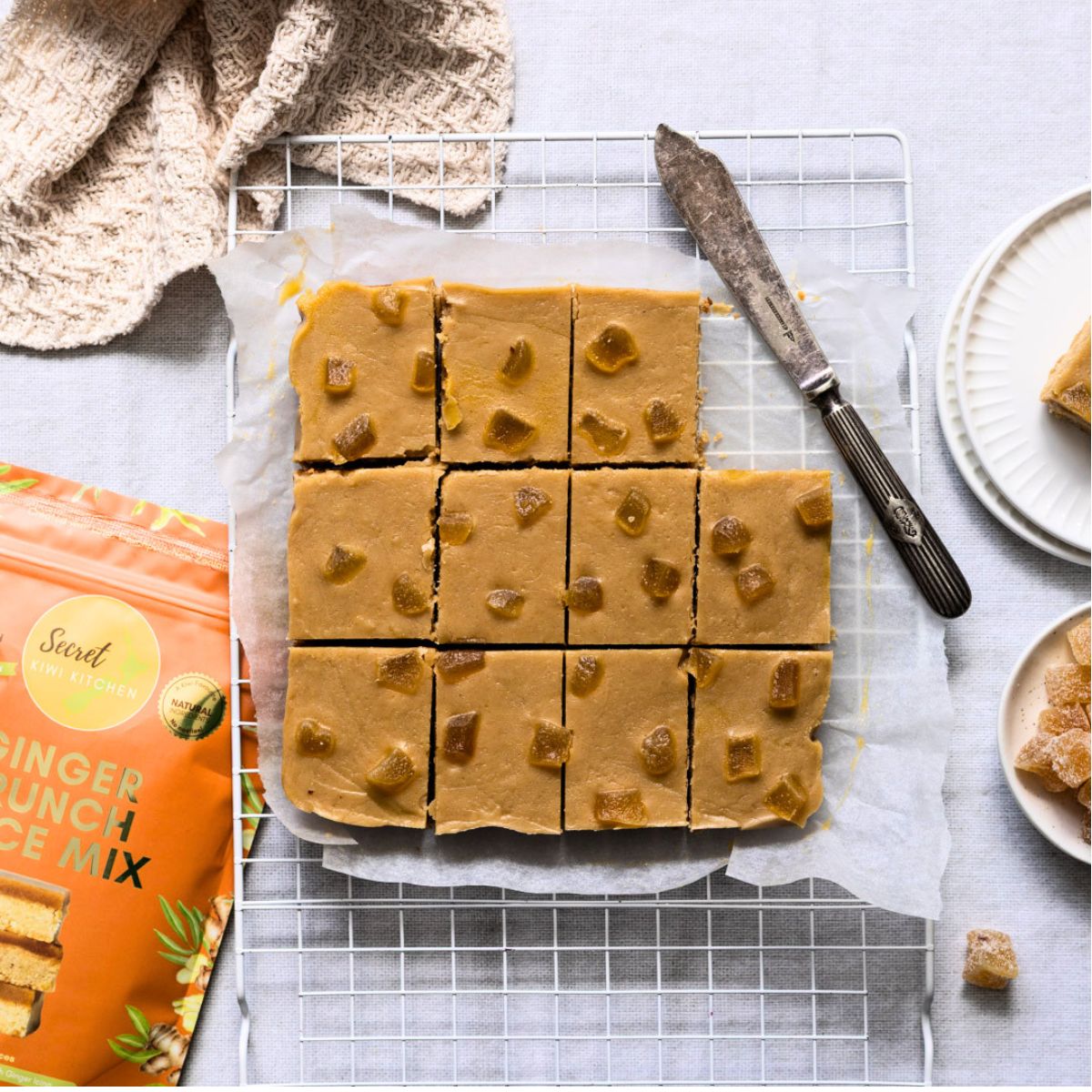 Secret Kiwi Kitchen Ginger Crunch Slice bars arranged on a wire cooling rack, with a knife, cloth, and packaged ginger punch mix visible around them on a light gray surface.