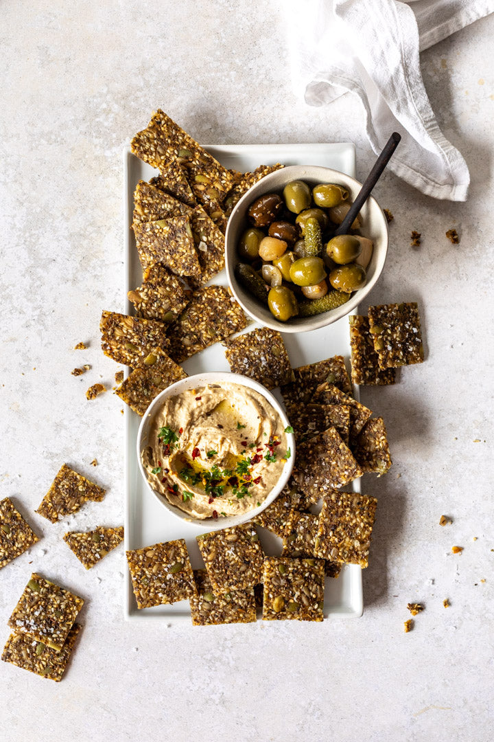 Secret Kiwi Kitchen Seeded Cracker squares with two small bowls of hummus and olives on a light surface.
