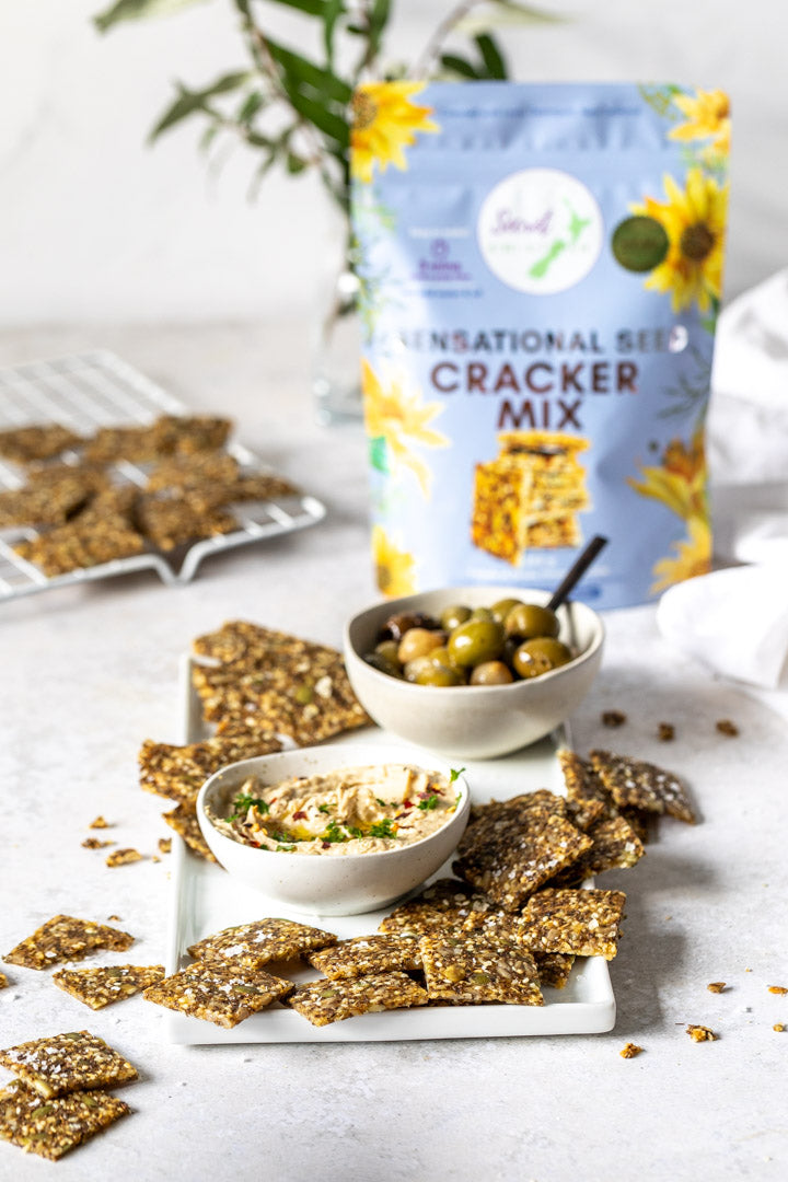 Secret Kiwi Kitchen Sensational Seed Cracker Mix packaging with crackers and snacks on a table