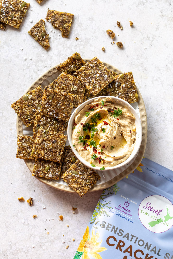 Platter of crackers with a bowl of hummus on a light surface, featuring Secret Kiwi Kitchen packaging.