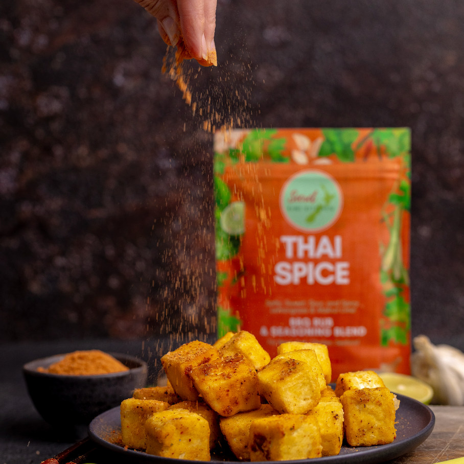 Person seasoning tofu cubes with a sprinkle of Secret Kiwi Kitchen Thai spice packet in the background